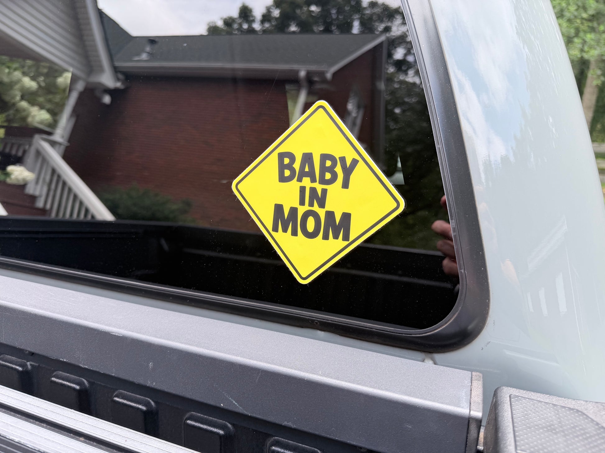 Baby In Mom™ vehicle decal: A yellow diamond-shaped vinyl sticker with the text 'BABY IN MOM' in black lettering, shown applied to the window of a light-colored pickup truck outdoors.