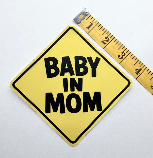 Baby In Mom™ vehicle decal: A yellow diamond-shaped sign with black border and the text 'BABY IN MOM' in bold black letters, shown on a white background next to a measuring tape.